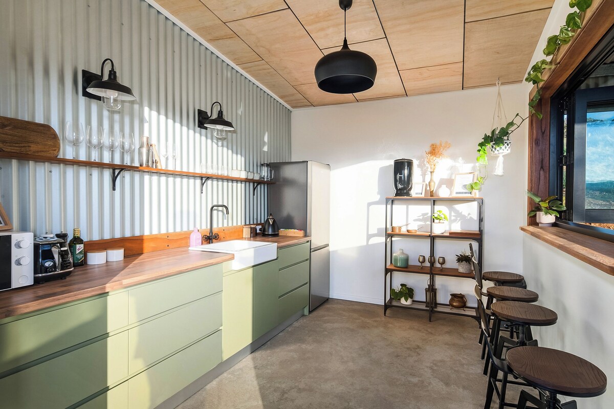 A modern kitchen with a wooden countertop, green cabinets, and a built-in sink. There is a bar area with stools and a shelf with decorative items.
