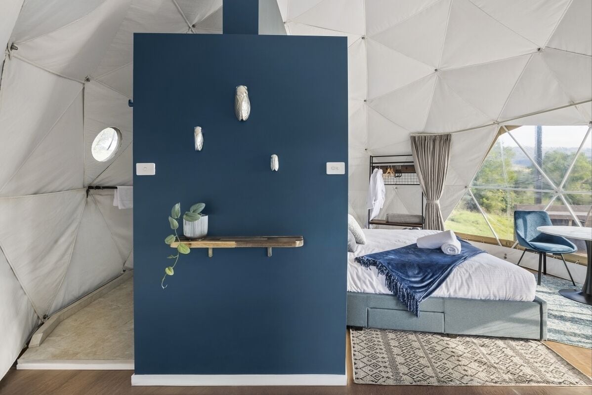 Una habitación moderna tipo tienda de campaña con una cama, un pequeño estante con plantas y vistas al exterior.