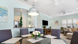 Un coin repas avec une table en verre, des sets de table blancs et un vase rempli de fleurs blanches. Des chaises violettes sont disposées autour de la table, et un ventilateur de plafond est installé au-dessus.