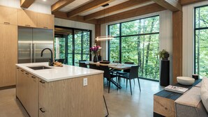 A modern kitchen with a central island, wooden beams, large windows, and a dining area with a view of the forest.