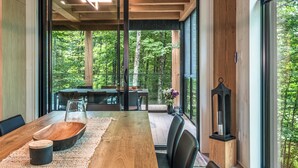 A modern dining area with a wooden table, black chairs, and a glass-top table. Large windows offer a view of a forested area.