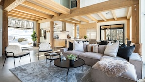 A modern interior with a high ceiling, wooden beams, a cozy seating area with a sofa and chairs, a kitchen with stainless steel appliances, and large windows offering a view of the outdoors.