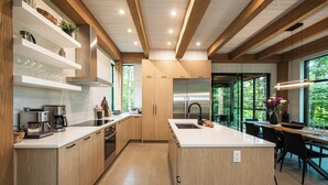 A modern kitchen with wooden cabinets, stainless steel appliances, and large windows offering a view of the outdoors.