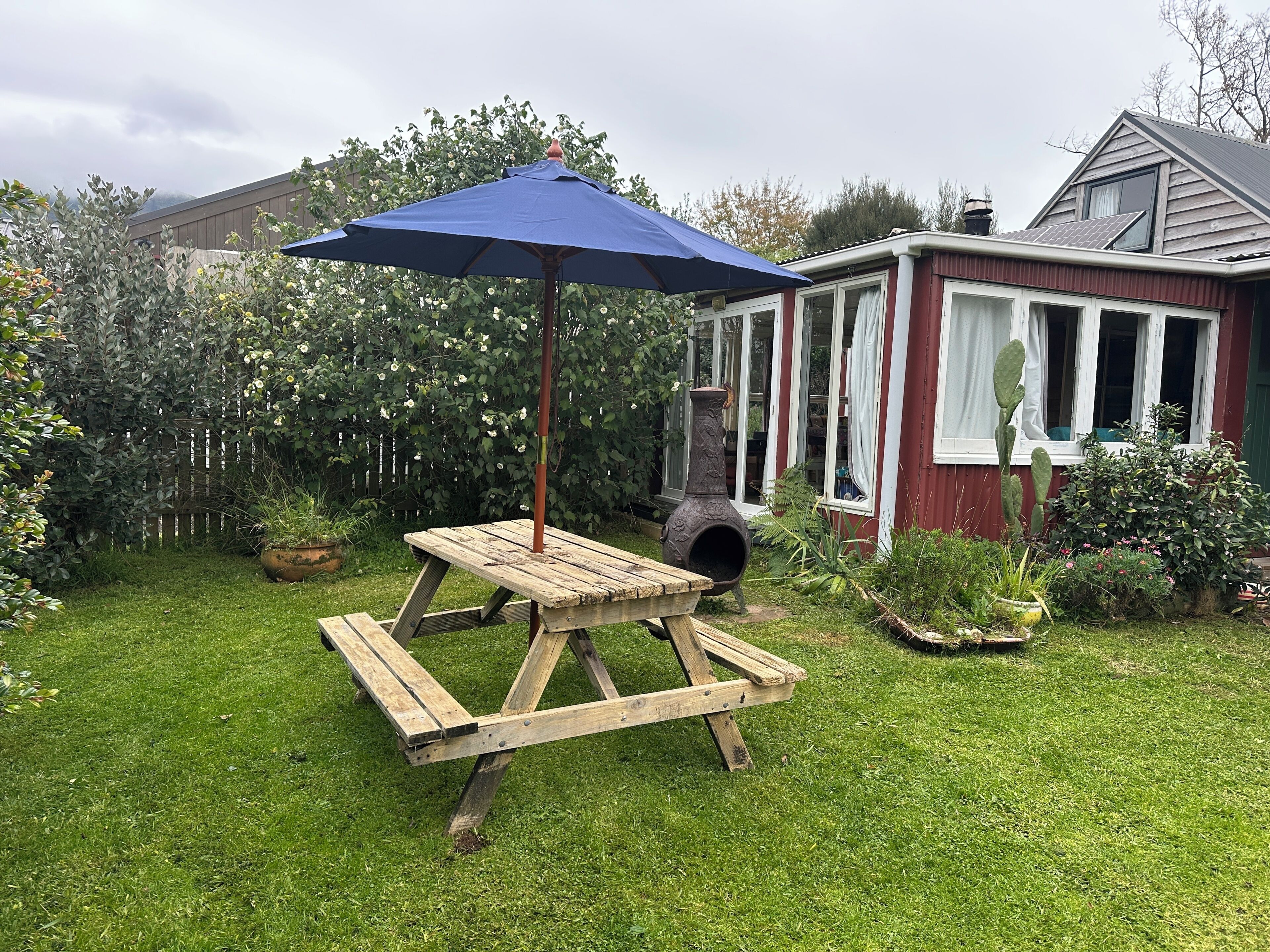 Una mesa de picnic de madera con un paraguas azul, rodeada por un jardín con diversas plantas y una casa al fondo.