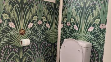 Ein Badezimmer mit Toilette, einem Fenster mit dekorativem Gitter und einer Wand mit floralem Tapetenmuster.