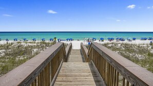 A wooden boardwalk leading to a beach with blue umbrellas and clear skies.