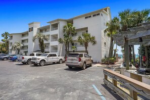 Un parcheggio con diversi veicoli, un edificio con numerosi balconi, palme e un pergolato in legno.