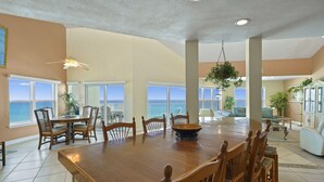 A spacious dining area with a large wooden table, surrounded by chairs, and large windows offering a view of the ocean.