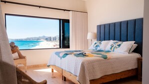 A bedroom with a large window offering a view of the beach and ocean, a bed with a blue headboard, and a wicker chair. - Casa Del Mar Las Olas Beachfront 2BR Penthouse (San Jose del Cabo)