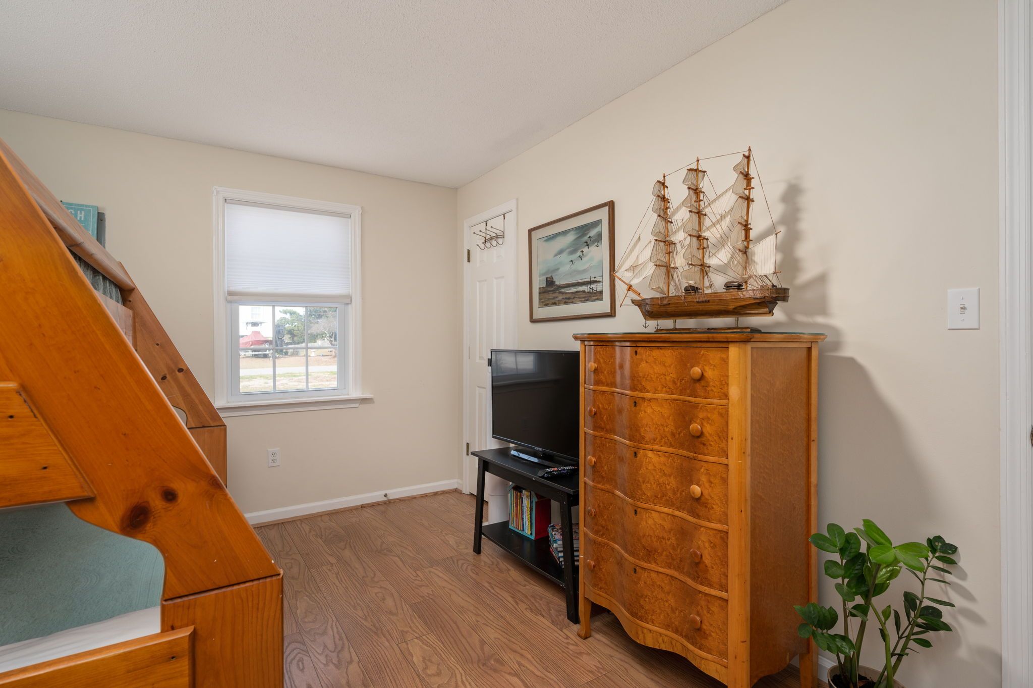 Un salon avec un escalier en bois, un téléviseur posé sur un meuble, une commode en bois avec des tiroirs, un tableau encadré accroché au mur et une fenêtre avec des stores.