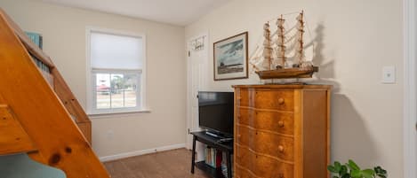 Un salon avec un escalier en bois, un téléviseur posé sur un meuble, une commode en bois avec des tiroirs, un tableau encadré accroché au mur et une fenêtre avec des stores.