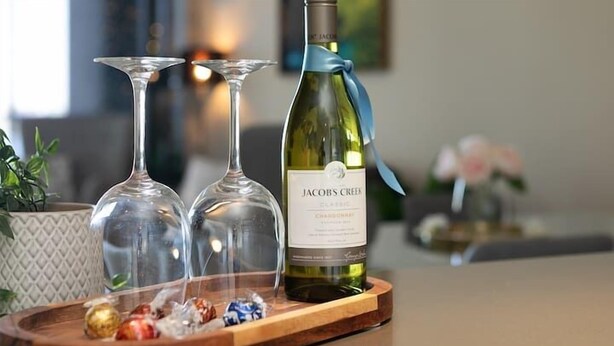 Une bouteille de Chardonnay Jacob's Creek sur un plateau en bois, accompagnée de trois verres à vin et de quelques amuse-gueules.