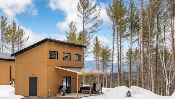 Une cabane en bois moderne dotée d’un patio couvert, entourée de neige et d’arbres.