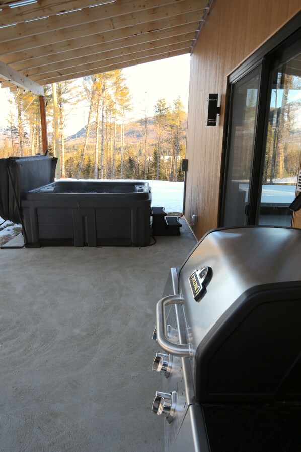 A covered outdoor area with a hot tub, a grill, and a view of a snowy landscape.