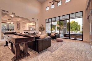 A spacious living area with a pool table, comfortable seating, and large windows offering a view of the outdoors.