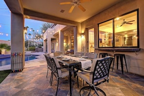 An outdoor patio with a dining table set for four, surrounded by chairs, and a pool in the background.