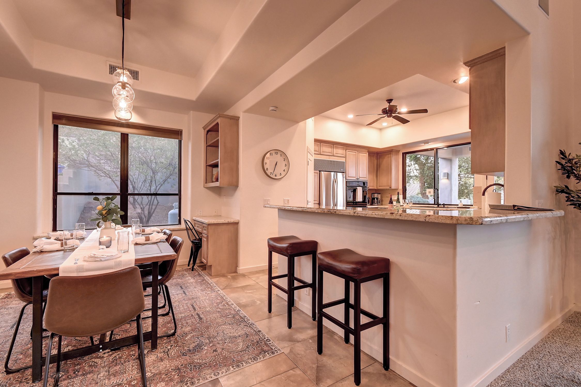 A modern kitchen with a central island, bar stools, a dining area with a table set for four, and a large window providing a view of trees.