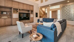 A spacious living room with a large flat-screen TV, a blue sofa, a white armchair, and a coffee table.