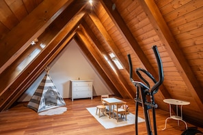A cozy attic room with a sloped ceiling, a small white dresser, a dining table with chairs, a tent-like play area, and a black exercise bike.