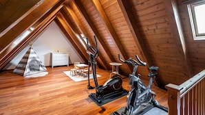 Um quarto de sótão com piso de madeira, teto inclinado, um aparador branco, uma mesinha e duas bicicletas ergométricas, além de uma tenda.