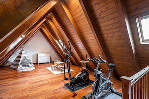 A wooden attic room with a sloped ceiling, wooden flooring, a white dresser, a small table, two exercise bikes, and a tent. - Cuveshic by SeaMount Rentals (Bossòst)