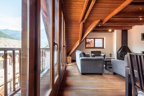 Ein gemütliches Loftzimmer mit einem großen Schiebeglastür, die auf einen Balkon führt, Holzbalken an der Decke, einer Couch, einem Tisch und einem Kamin.