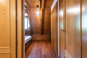Ein schmaler Gang mit Holzwänden und schräger Decke, der in ein Schlafzimmer mit Bett und Pendelleuchte führt.