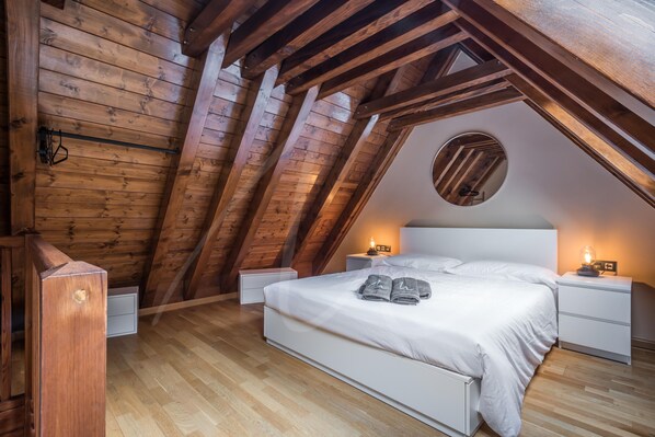 A bedroom with a sloped ceiling, wooden beams, a round skylight, a bed with white linens, two bedside tables with lamps, and a wooden wardrobe.