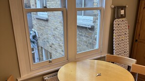 A room with a round wooden table and four chairs, large windows with a view of a brick building, and a radiator under the windows. - Quiet Paddington studio Apartment (London)