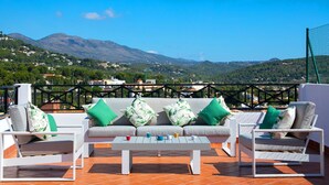 A terrace with a sofa and chairs, a table, and a view of a mountainous landscape.