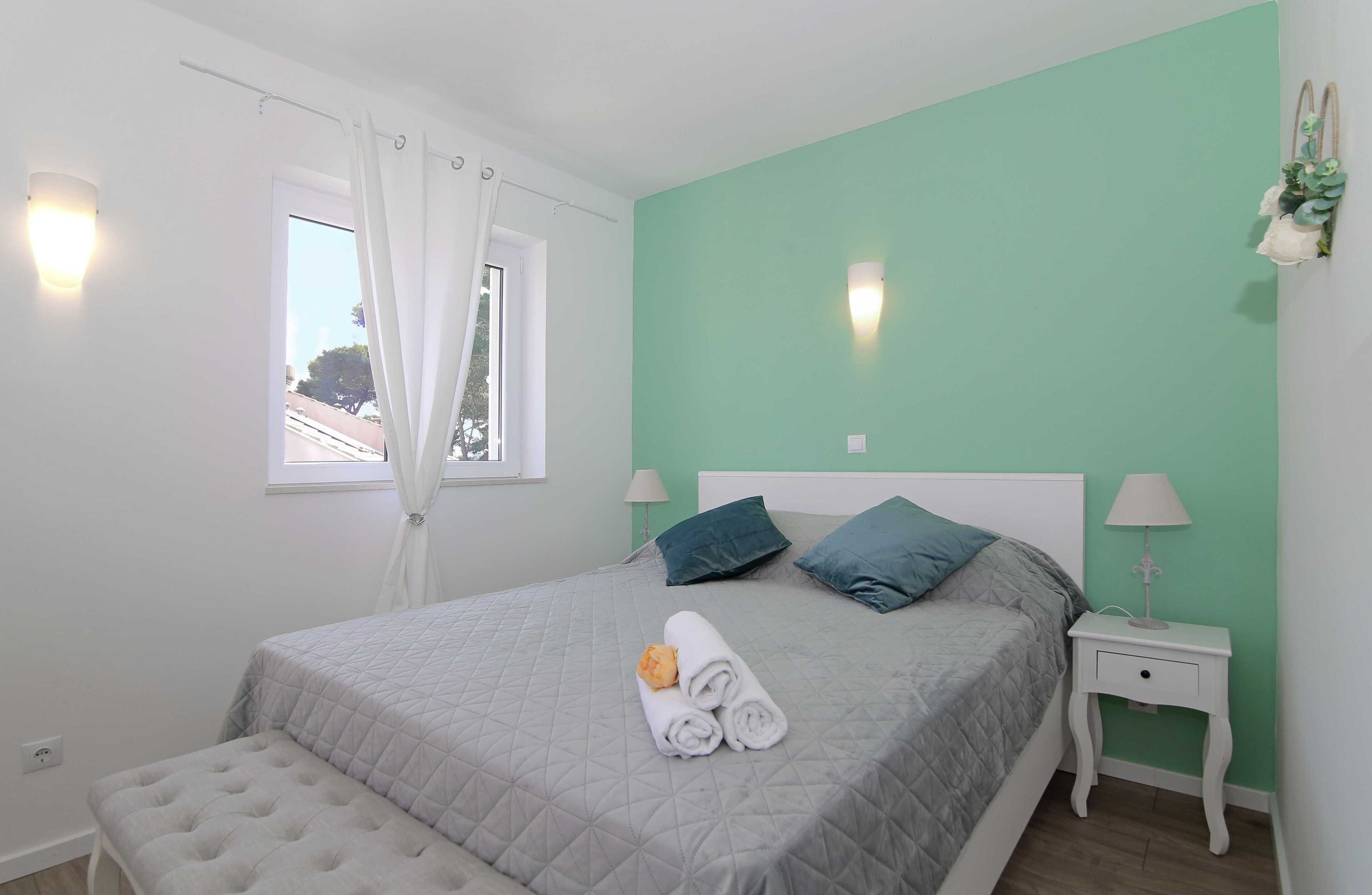 A neatly made bed with pillows, a bedside table with a lamp, and a window with curtains.