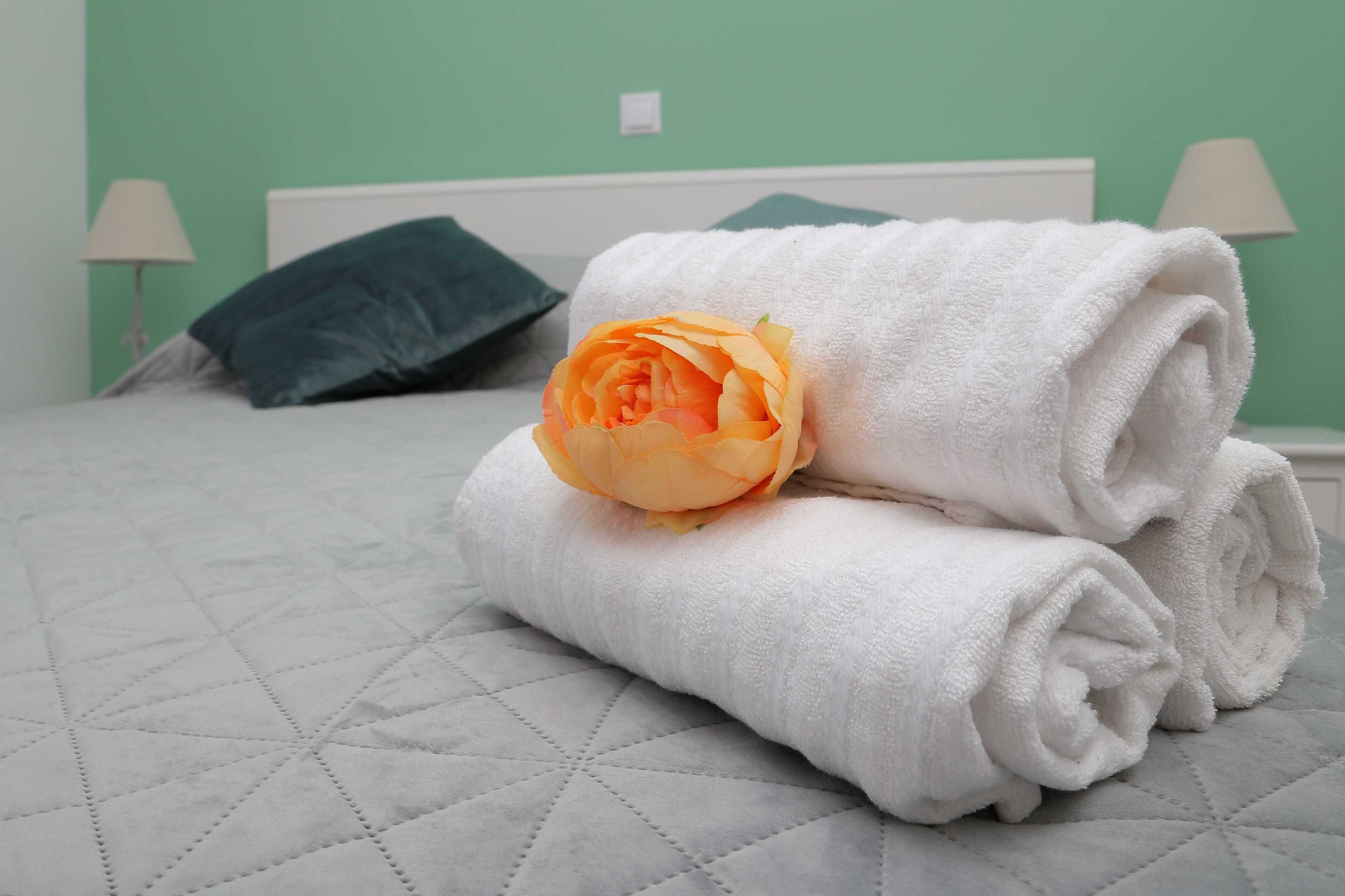 Una cama bien hecha con una colcha acolchada, una toalla blanca enrollada y una sola flor naranja.