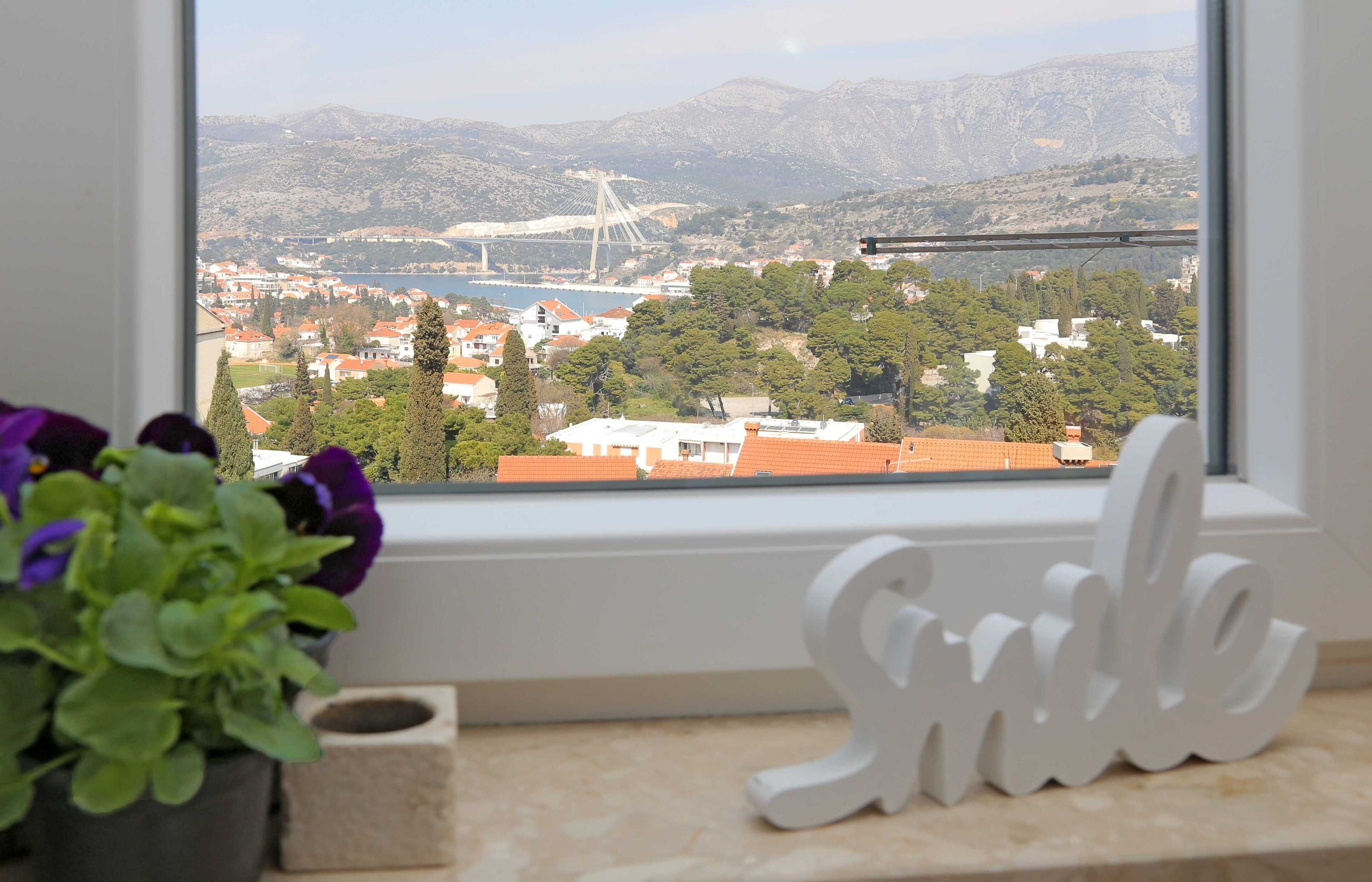 A window view of a coastal town with a bridge, mountains, and a 'Smile' decoration.
