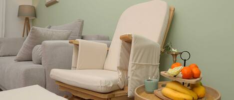 Una habitación con un sofá gris, una silla de madera con cojín blanco, una mesita redonda con frutas y una vela, y una lámpara sobre un atril.