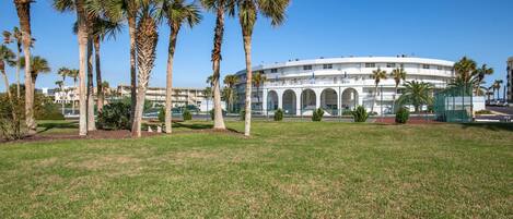 A large, white, multi-story building with a curved roof and arched windows, surrounded by a well-maintained lawn and palm trees.