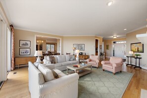 A spacious living room with a large sofa, a coffee table, and two armchairs. There is a dining area in the background with a chandelier.