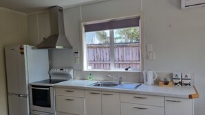A compact kitchen with white cabinets, a built-in oven, a stove, a sink, and a window with a view of a wooden fence. - The Pink House (Tokoroa)