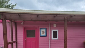 A pink building with a wooden porch, a red door, and a small window. - The Pink House (Tokoroa)