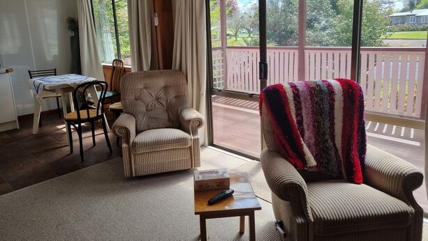 A living room with a sofa, armchair, and a table with a remote control. There is a window with curtains and a view of a balcony. - The Pink House (Tokoroa)