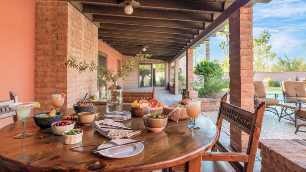 A covered patio with a wooden table set for a meal, surrounded by brick walls and outdoor furniture. - Charming 5BDR Hacienda w/ Pool, Fire Pit, Billiards | La Casona by AvantStay (Phoenix)