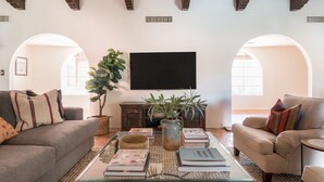A living room with a sofa, armchair, and coffee table, featuring books, a plant, and a television. - Charming 5BDR Hacienda w/ Pool, Fire Pit, Billiards | La Casona by AvantStay (Phoenix)