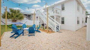 A white building with a balcony and a staircase leading to the upper floor. There are blue Adirondack chairs and a small table with a striped umbrella.