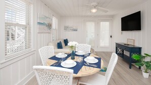 A dining area with a round table set for four, white wicker chairs, a ceiling fan, and a TV mounted on the wall.