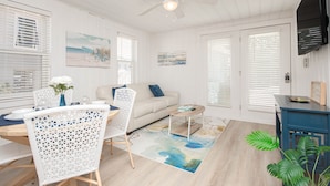 A bright, white-themed living room with a dining area, a ceiling fan, and a large window with blinds.