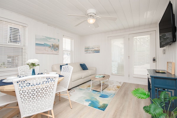 A bright, white-themed living room with a dining area, a ceiling fan, and a large window with blinds. - Stepstobeach*BigDogFriendly*Outdr Shower (North Myrtle Beach)