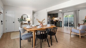Un comedor con una mesa de madera dispuesta para cuatro personas, rodeada de sillas negras. Hay una mesita auxiliar con una lámpara y un jarrón con flores secas. Un espejo grande cuelga en la pared sobre la mesa.