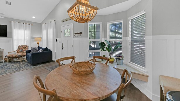 A spacious living room with a wooden dining table, comfortable seating, a large window with blinds, and a hanging light fixture. - A Sunshine Daydream | 30A Home w/ Community Pool + Outdoor Shower (Santa Rosa Beach)