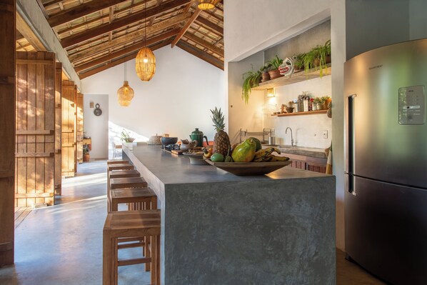 A kitchen with a large island, wooden stools, a stainless steel refrigerator, and potted plants on shelves. - Casa Canoa (Maraú)