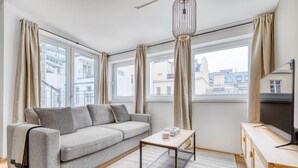 Una sala de estar moderna con un sofá gris, una mesita pequeña y un televisor sobre un mueble de madera. Amplias ventanas ofrecen vistas a la ciudad.