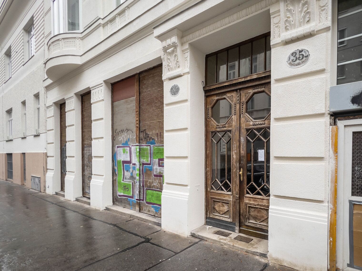 A building with a white facade, a closed wooden shutter door, and a large glass window with a number '35' on the right.